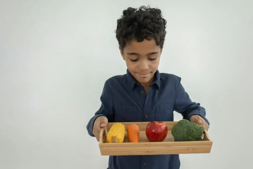 Smiling boy holding a tray of fresh fruits and vegetables, promoting healthy eating.